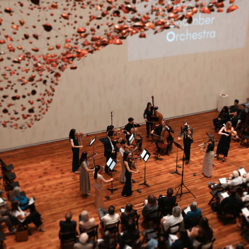 An orchestra performs on a wooden stage in front of an audience, with a large, abstract art installation hanging overhead.
