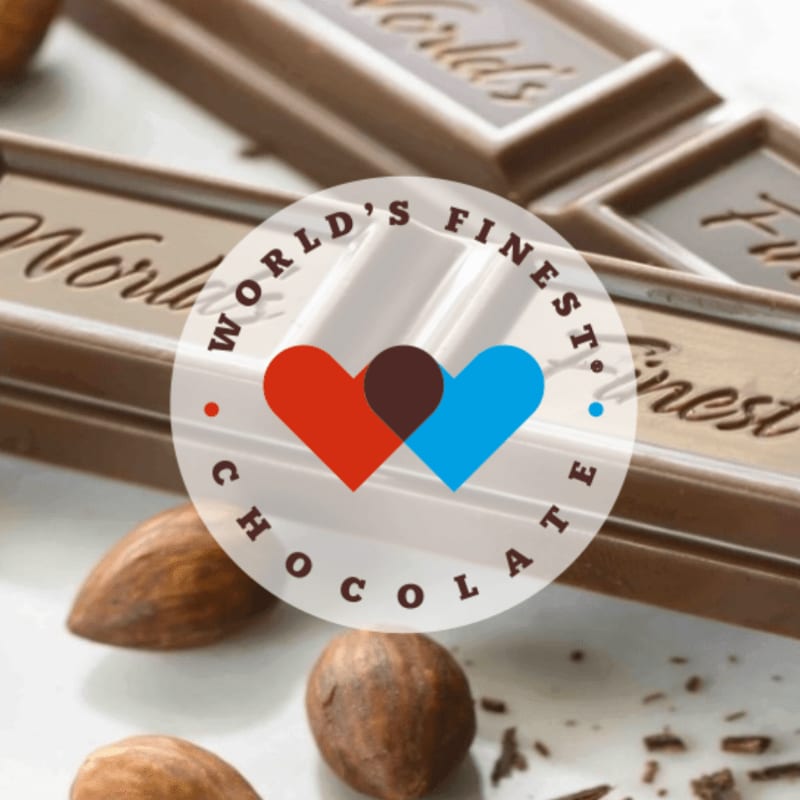 Several almonds and chocolate pieces are in the foreground, with a logo overlaying a blurred background of chocolate bars.