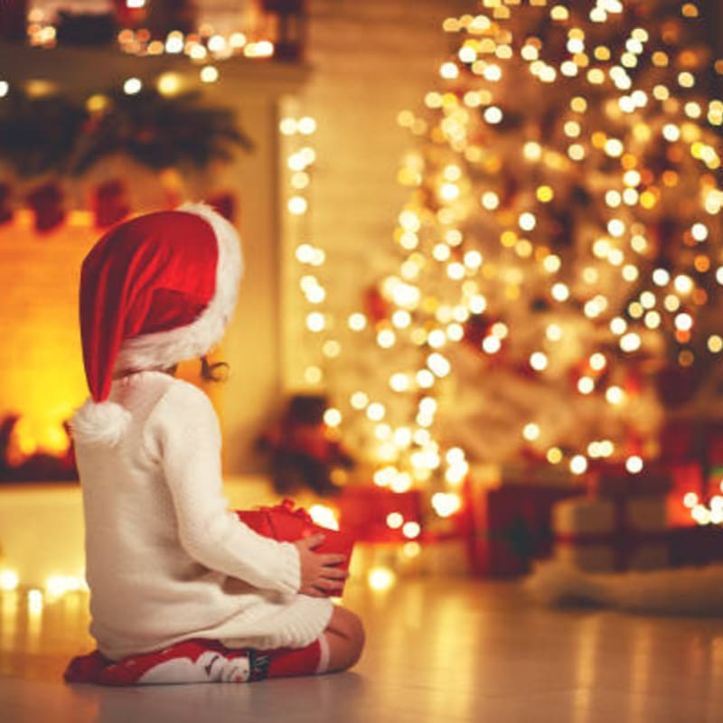 A child in a Santa hat sits on the floor holding a gift, with a brightly lit Christmas tree and fireplace in the background.