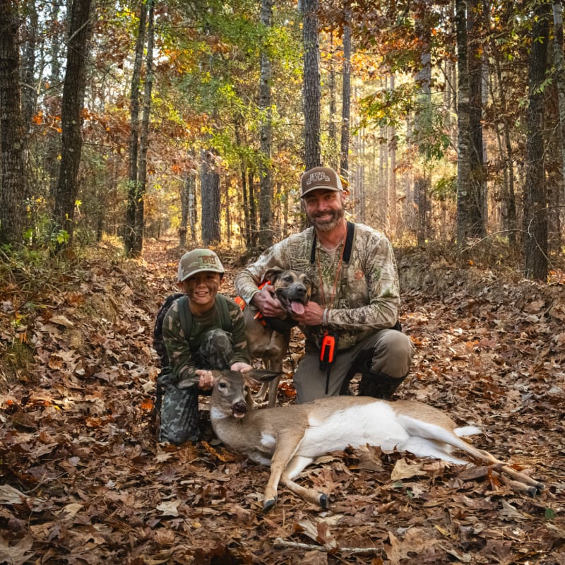 A man and a boy, with a dog, pose with a harvested deer in the leaf-strewn foreground of a sun-dappled autumn forest.
