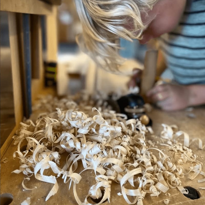A child with blonde hair is planing wood, creating a pile of shavings in the foreground, with a blurred background of a workshop.
