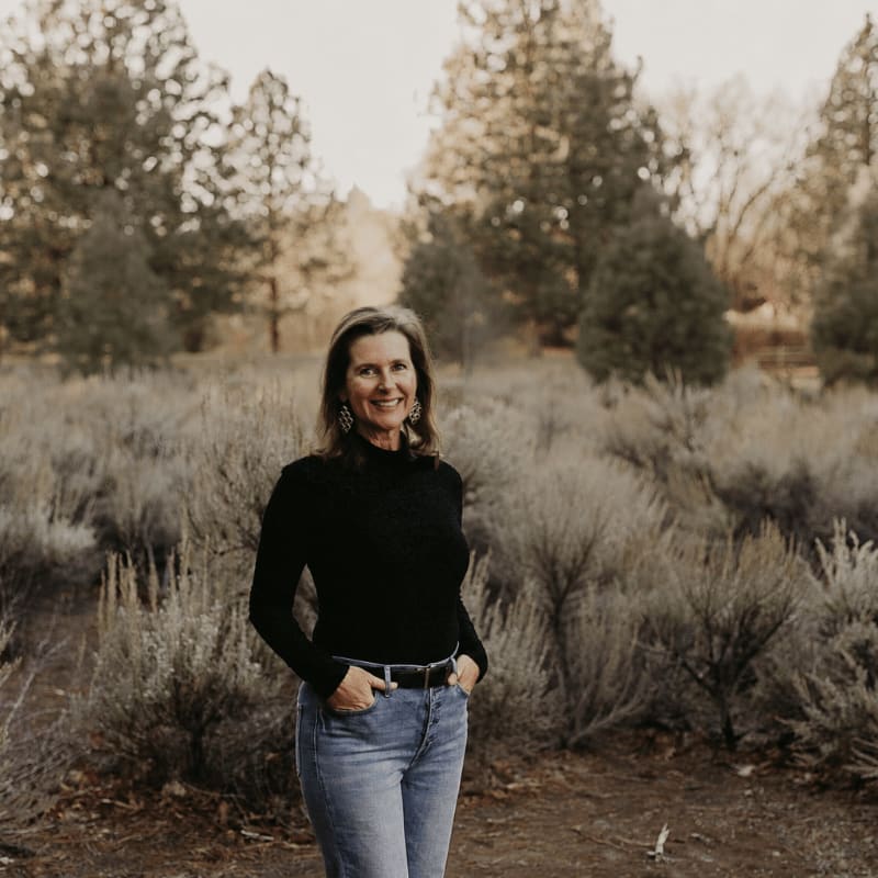 A woman in a black turtleneck and jeans stands with her hands in her pockets in a field of dry brush, with trees and a soft sky in the background.