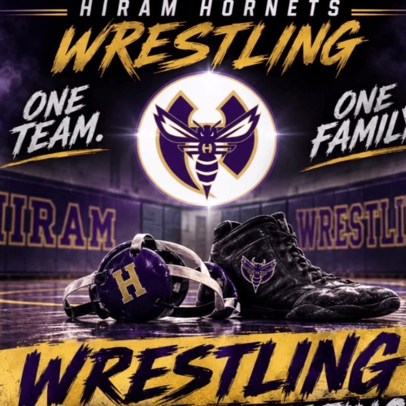 A wrestling shoe and headgear rest on a mat in front of a blurred background of a wrestling arena with the Hiram Hornets logo.