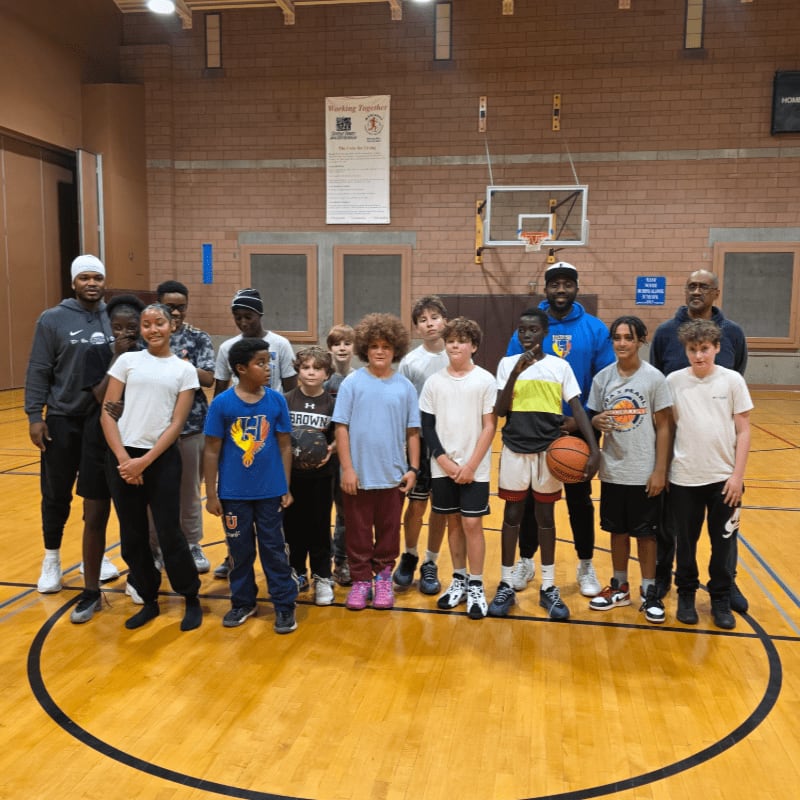 A group of young basketball players and coaches stand on a polished wooden court in front of a brick wall with a basketball hoop.