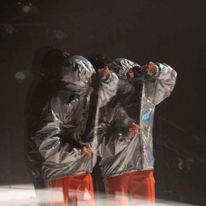 A person in a shimmering silver jacket and orange pants is captured in multiple overlapping frames against a dark, smoky background.
