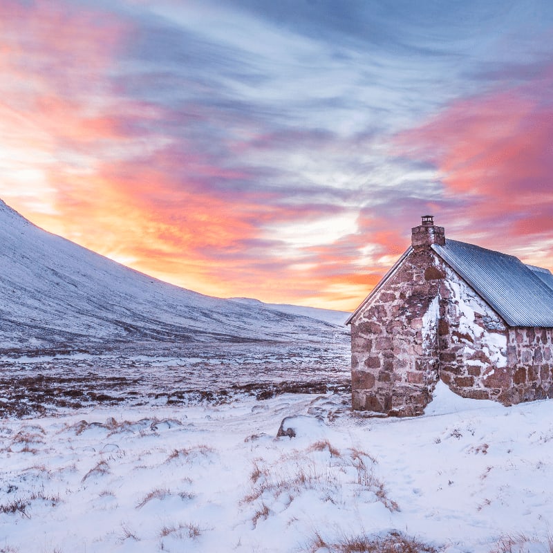 A stone cottage sits in a snowy landscape under a vibrant, colorful sunset sky.