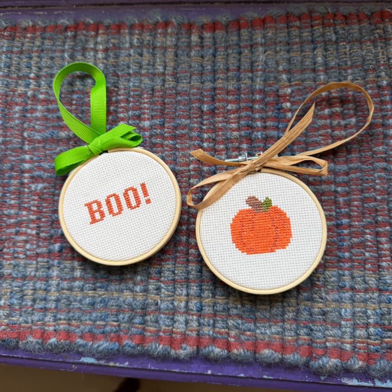 Two small, round cross-stitch ornaments, one with "BOO!" and the other a pumpkin, hang on a textured, woven surface.