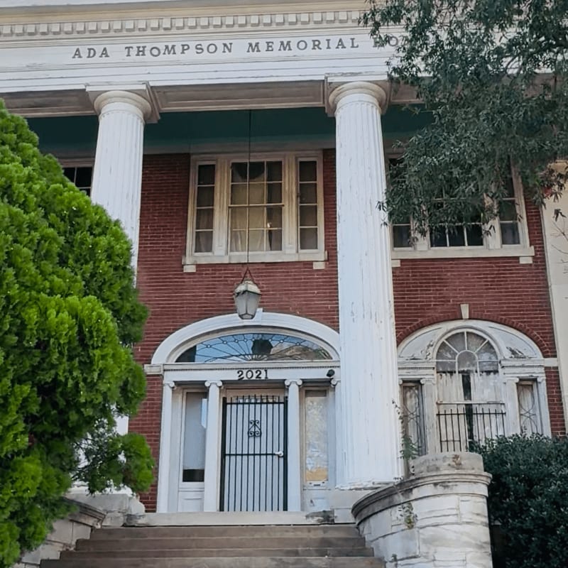 Lush green trees frame the weathered brick facade of the Ada Thompson Memorial building, with its grand columns and arched entryway.
