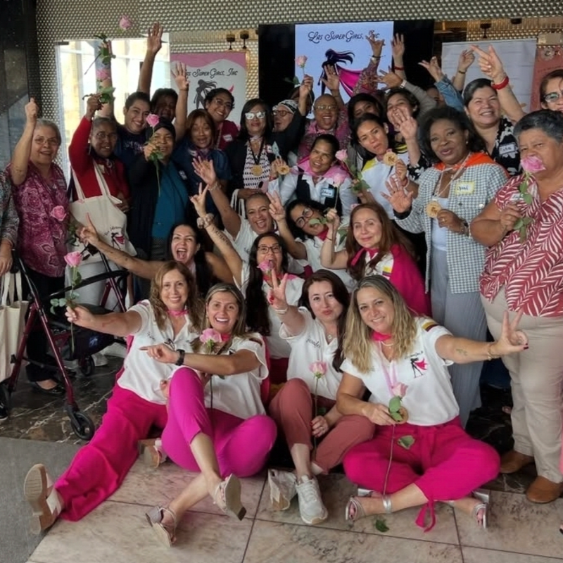 A group of women in pink and white pose for a photo, holding roses, with banners in the background.