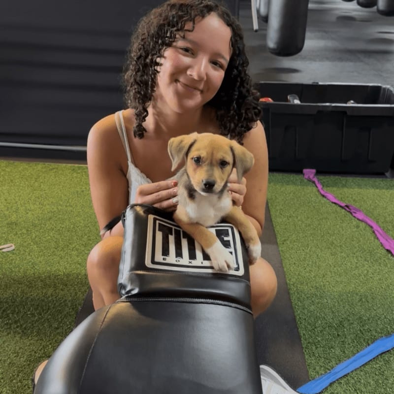 A young woman with curly hair smiles as she holds a small puppy on a boxing pad in a gym with punching bags in the background.