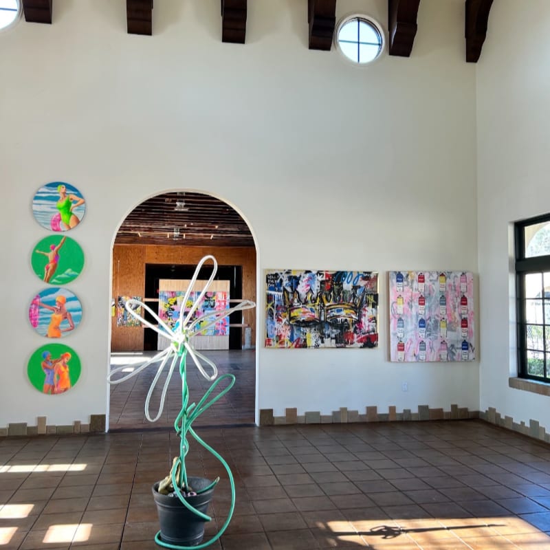 A green hose sculpture resembling a flower stands in the foreground of a brightly lit art gallery, with colorful paintings adorning the walls and an arched doorway leading to more exhibits in the background.