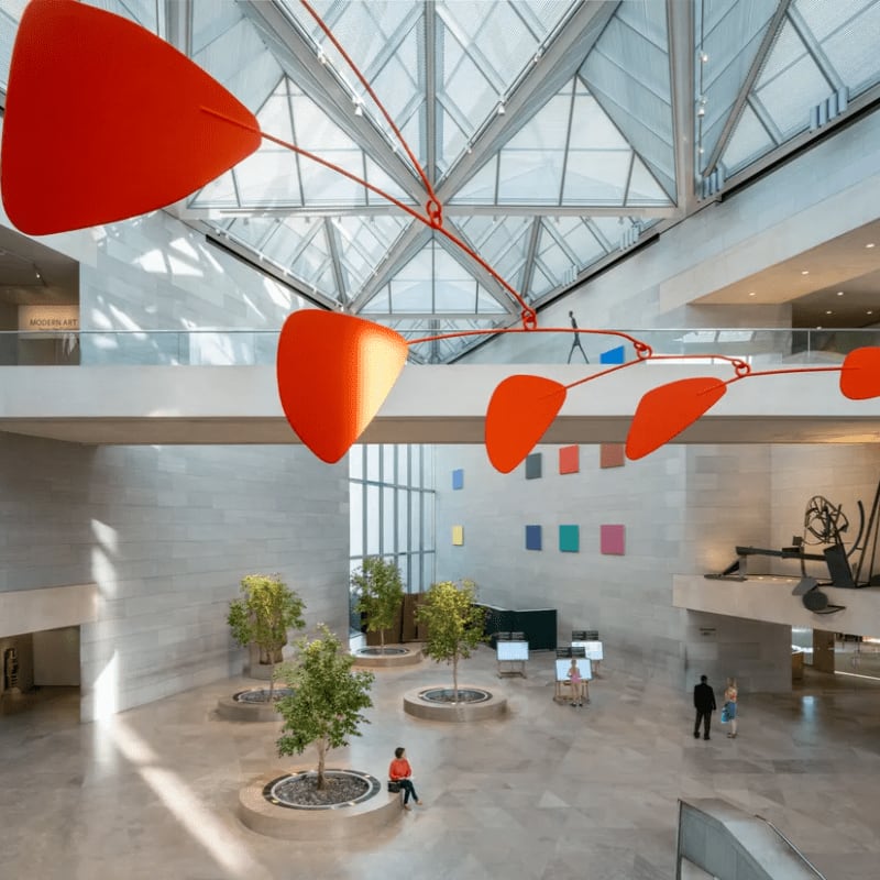A large orange mobile hangs in the foreground of a bright, modern museum atrium with trees and people scattered throughout the open space.