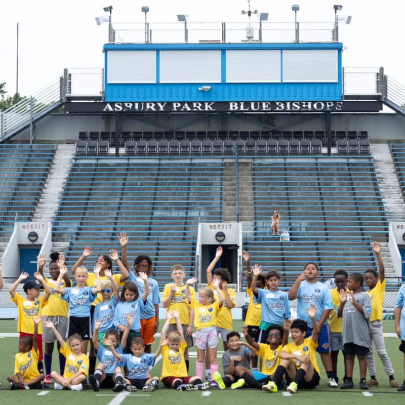 Children in colorful shirts wave from a soccer field in the foreground, with a large blue stadium and bleachers filling the background.