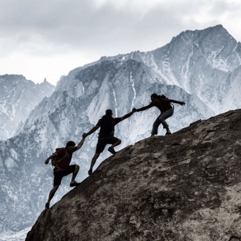 Three hikers ascend a rocky slope, reaching for each other, with a majestic mountain range looming in the background under a cloudy sky.
