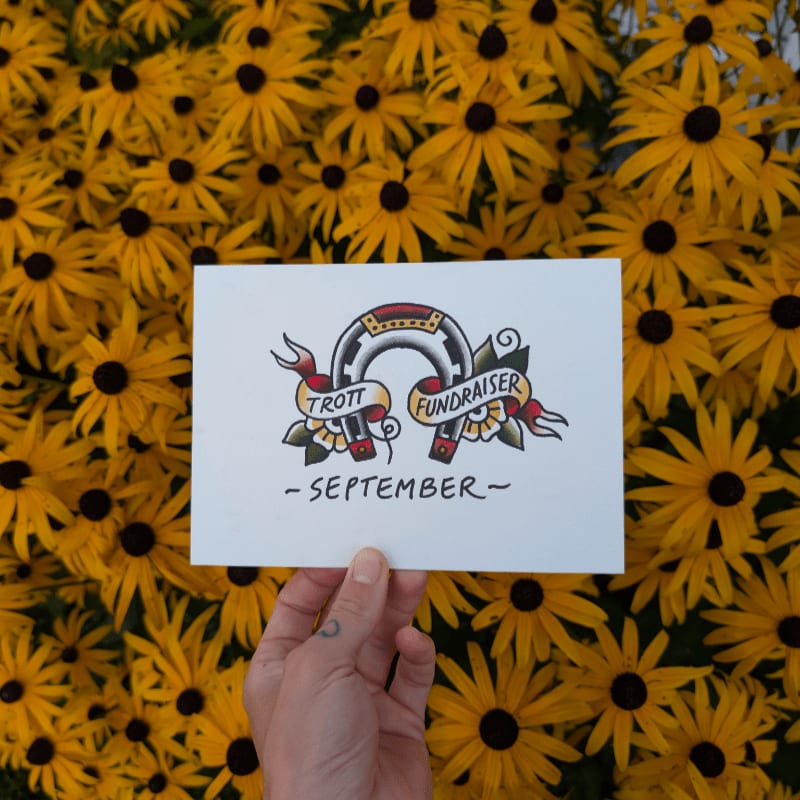 A hand holds a card with a horseshoe design and the words "Trott Fundraiser - September" against a vibrant background of yellow flowers.