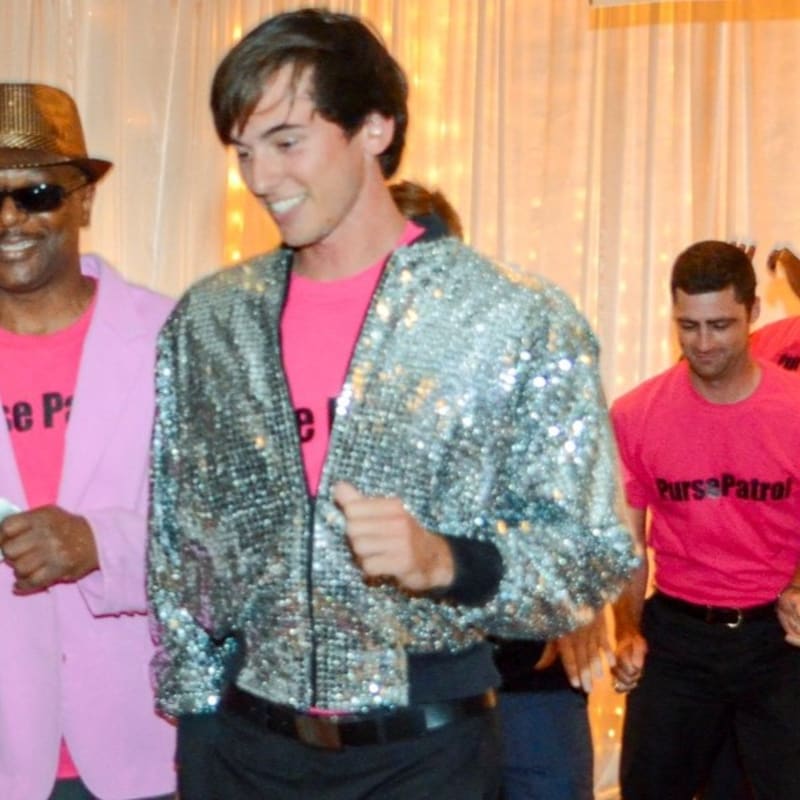 A man in a sequined jacket and pink shirt dances in the foreground, while other men in pink shirts and a man in a pink suit and fedora are visible in the blurred background.