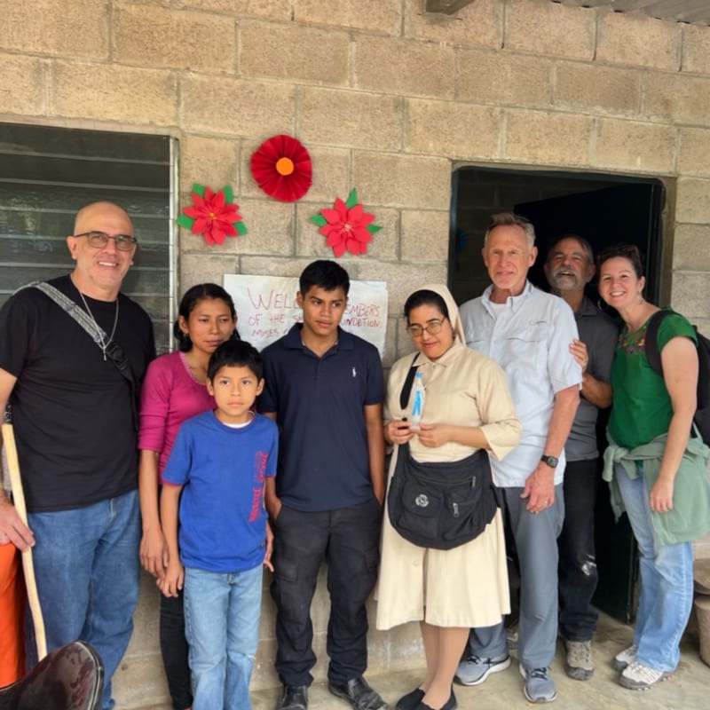 A group of people, including a nun and a young boy in the foreground, stand in front of a brick building decorated with red flowers.