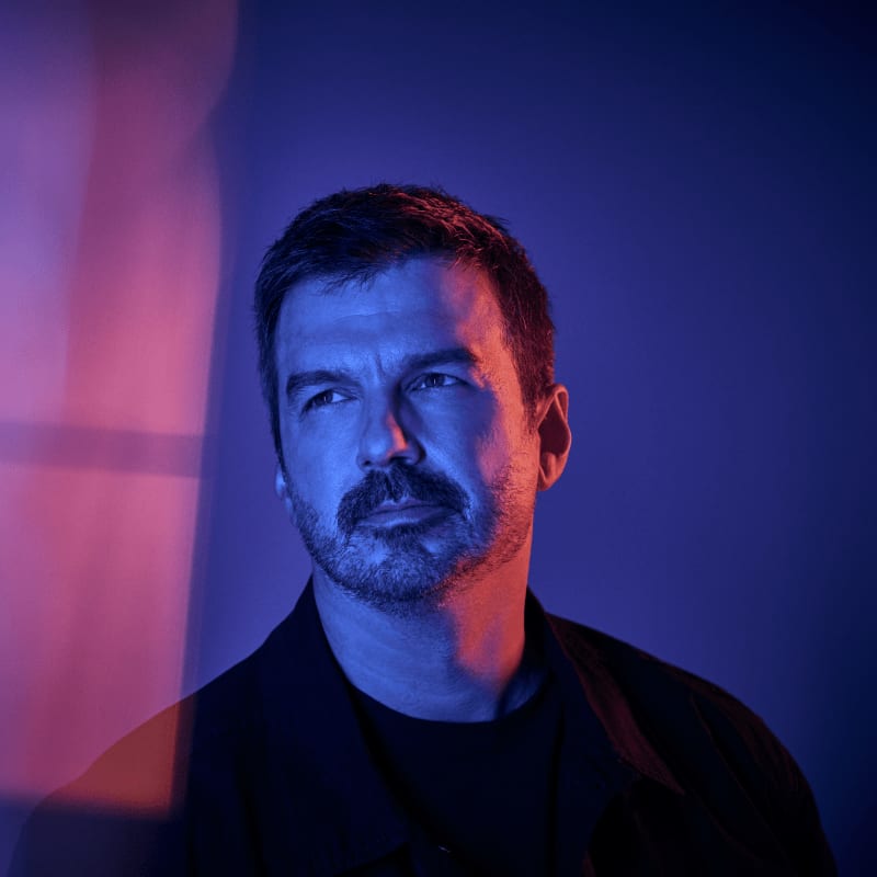 A man with a mustache looks off to the side, illuminated by pink and blue light against a dark background.