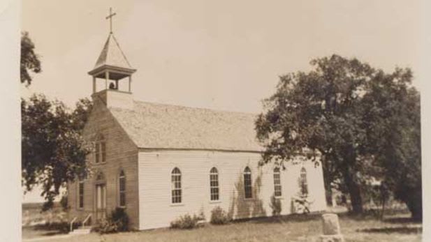 A white wooden church with a steeple stands in the foreground, surrounded by trees and a cloudy sky in the background.