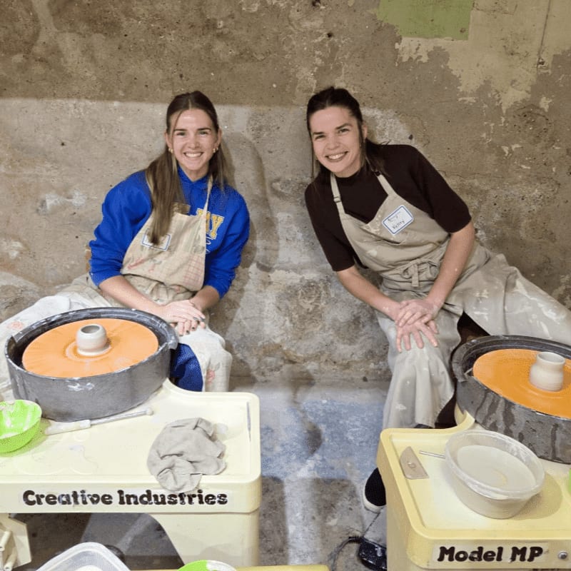 Two smiling young women in aprons sit at pottery wheels in a studio with a rough concrete wall in the background.