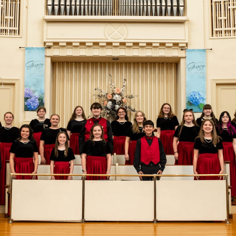 A choir of young people in red skirts and black tops stand in front of a floral arrangement and banners, with organ pipes visible in the background.