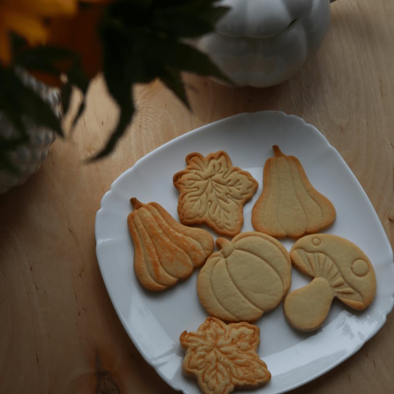 A white plate holds several autumn-themed cookies, including pumpkins, leaves, and a mushroom, with a blurred white pumpkin and yellow flowers in the background.
