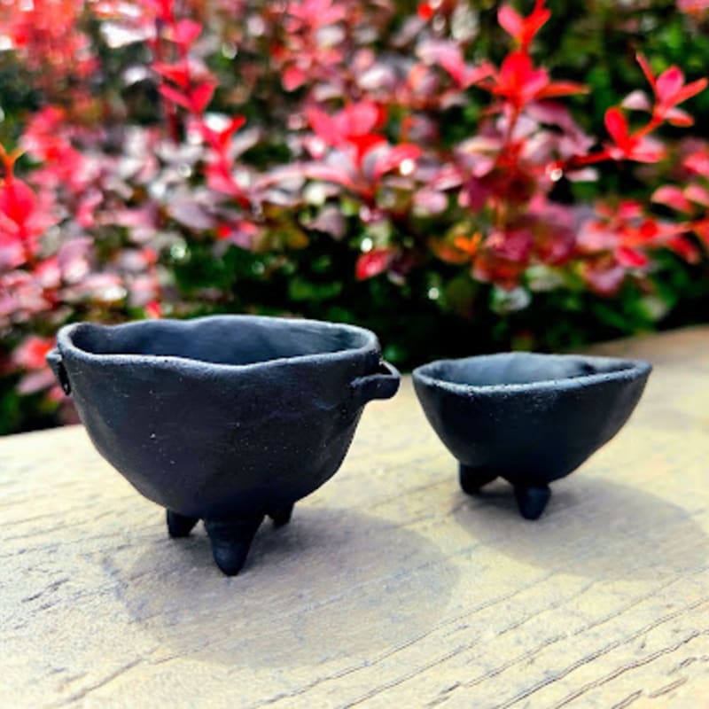 Two small, dark, three-legged pots sit on a textured surface in front of a blurred background of vibrant red foliage.