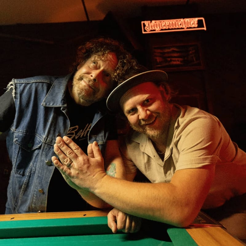 Two men pose by a pool table in the foreground, with a Jägermeister sign visible in the dimly lit background.