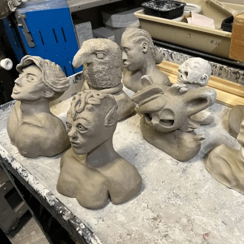 Several unfinished clay busts of fantastical creatures and humanoids are arranged on a workbench, with shelves and containers visible in the background.