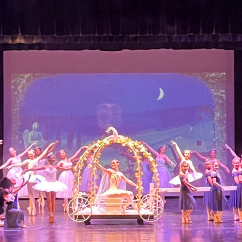 Dancers in tutus and costumes surround a Cinderella-like carriage on a stage with a starry night backdrop.