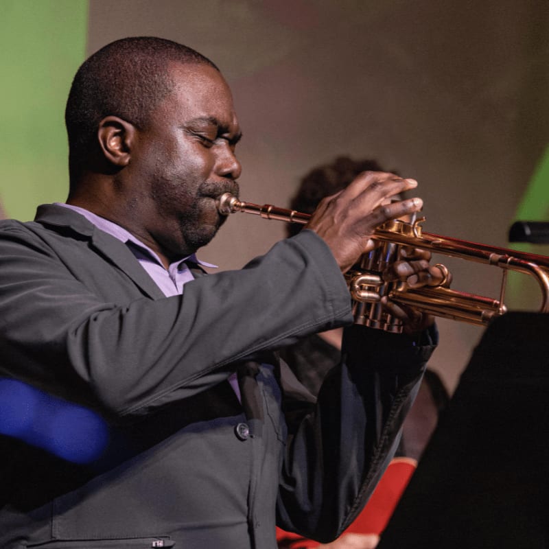 A man in a suit plays a trumpet in the foreground, with a blurred background of other musicians and colorful lighting.
