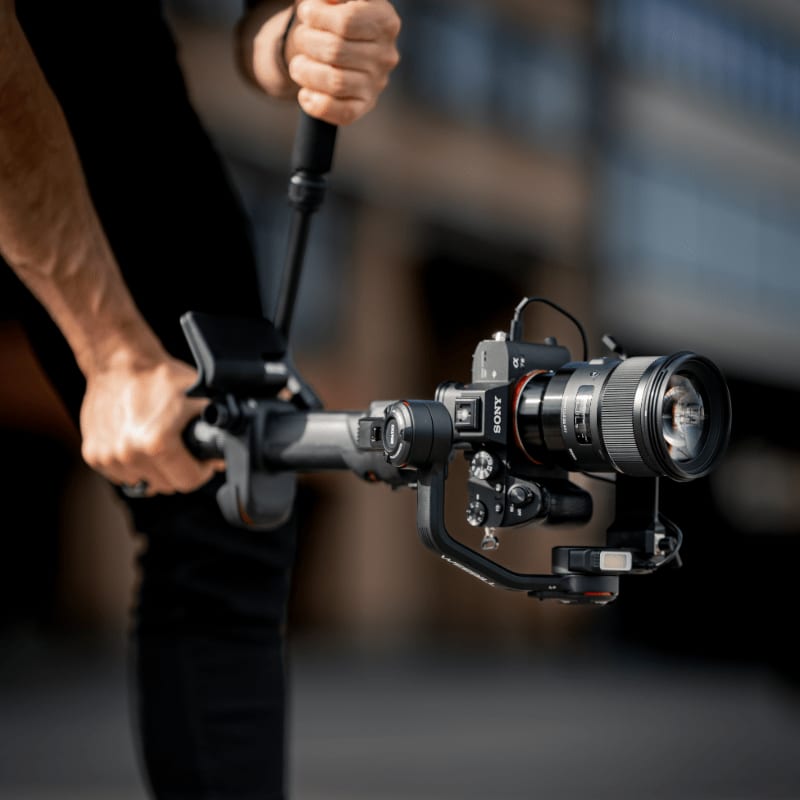Hands in the foreground hold a professional camera on a gimbal, with a blurred urban background.
