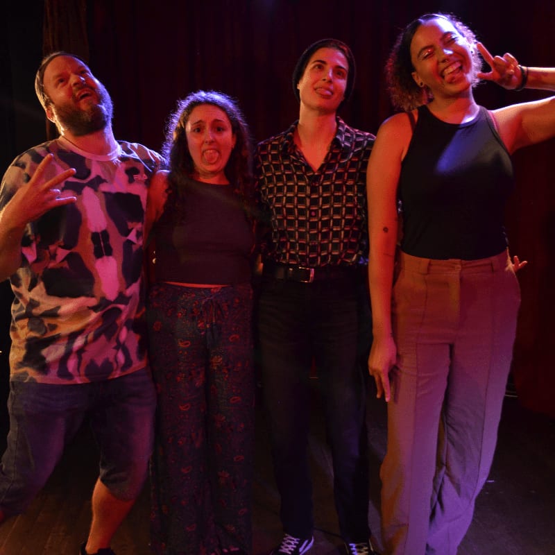 Four friends pose playfully in the foreground against a dimly lit, stage-like background.