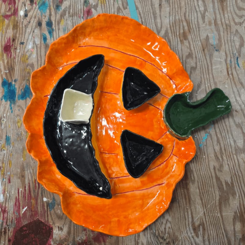 A ceramic pumpkin-shaped dish with black cut-out features and a green stem sits on a colorful, paint-splattered wooden surface.
