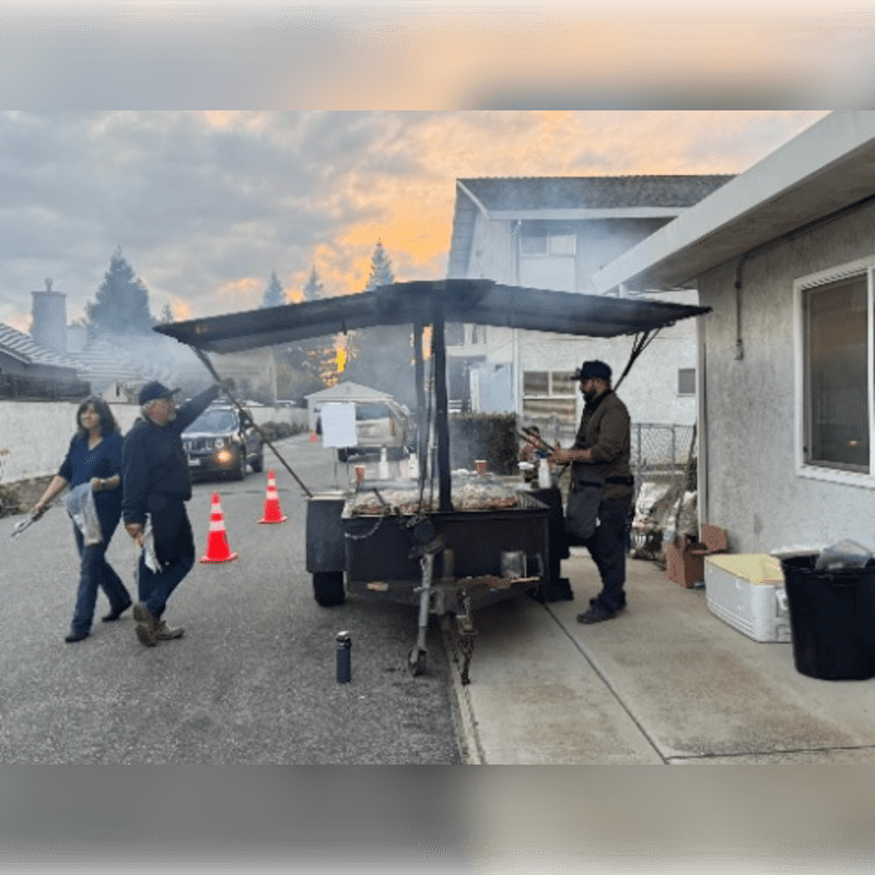 People gather around a smoking food trailer in a suburban driveway with houses and trees in the background under a cloudy sky.