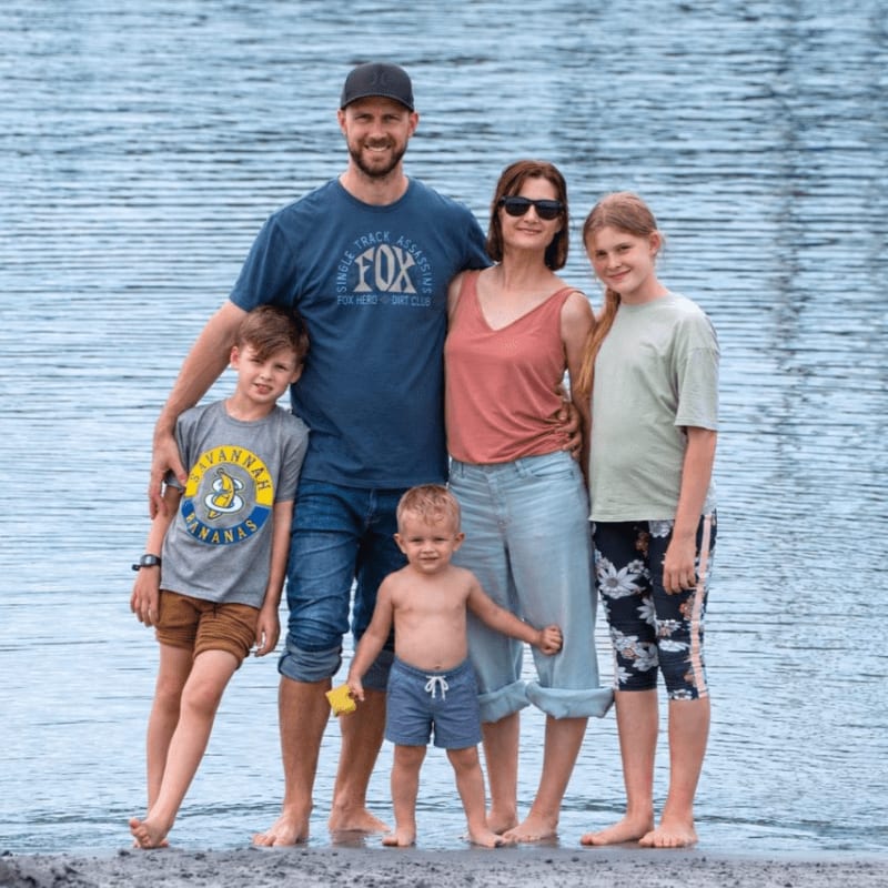 A family of five stands on a rocky shore with a calm blue body of water behind them.