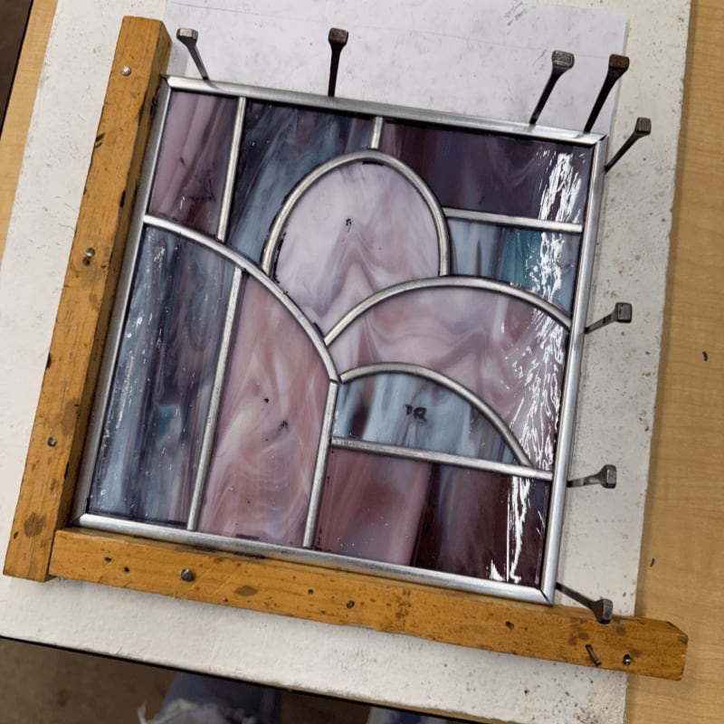 A stained glass panel with purple and blue hues is being held in place by a wooden jig with nails, resting on a white surface.
