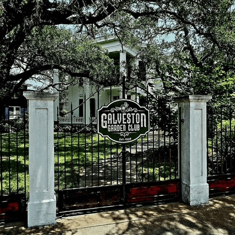 A black metal gate with a "Galveston Garden Club" sign stands in the foreground, leading to a white historic house with a porch and large trees in the background.