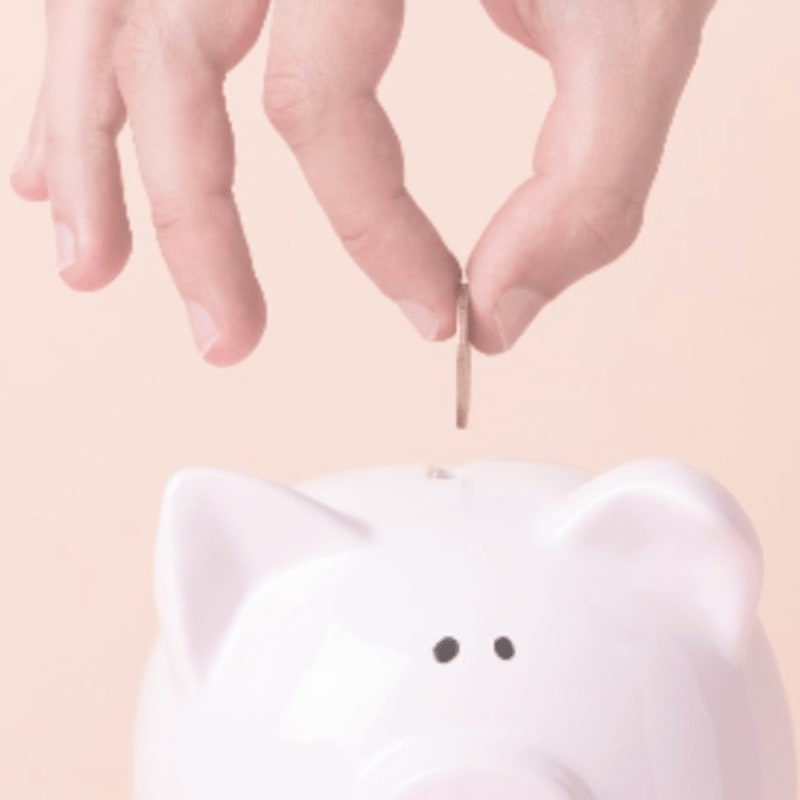 A hand drops a coin into a white piggy bank against a soft, peachy background.