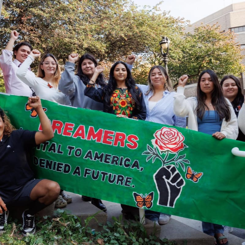 A group of young people raise their fists in solidarity behind a banner that reads "DREAMERS: VITAL TO AMERICA, DENIED A FUTURE," with a stylized fist holding a rose and butterflies, set against a backdrop of trees and a building.