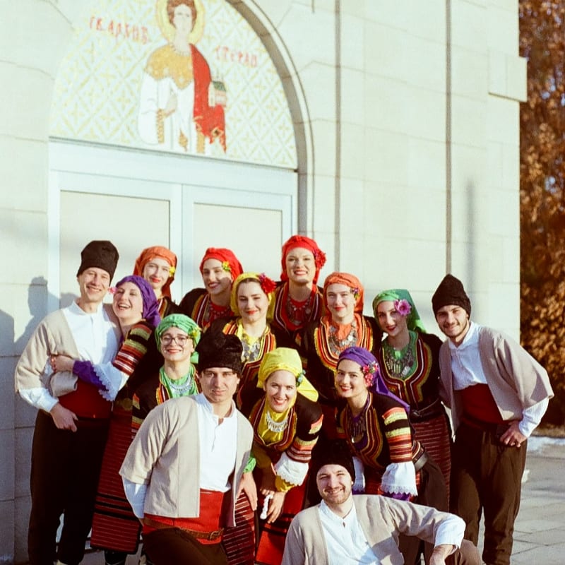 A group of people in traditional clothing pose in front of a building with a mosaic.