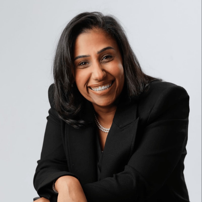 A smiling woman in a black suit leans on a chair against a plain gray background.