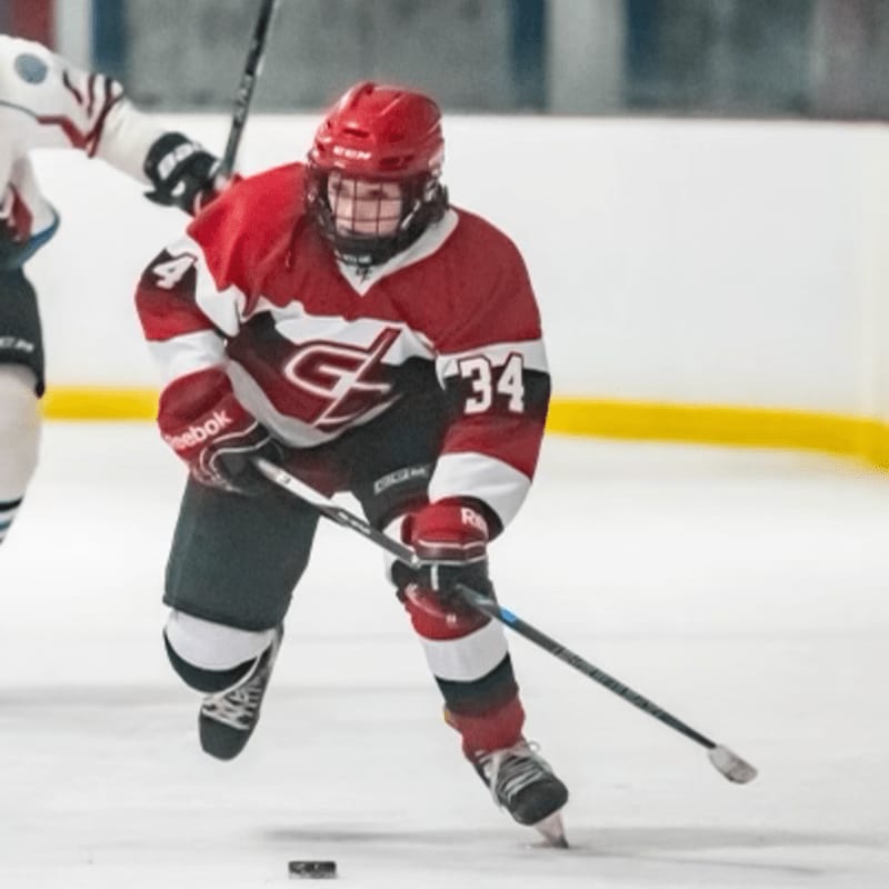A hockey player in a red jersey skates on an ice rink with a blurred background of another player and the boards.