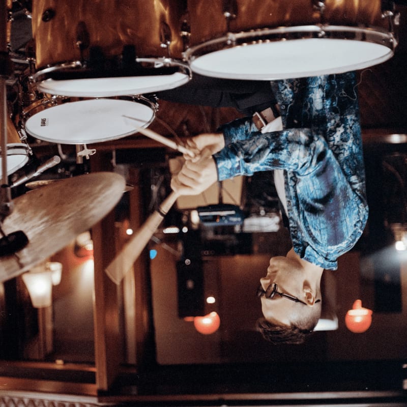 A person in a blue shirt plays drums in the foreground, with a blurred background of lights and wooden structures.
