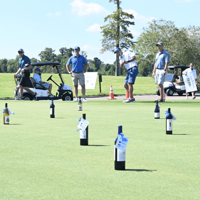 Several bottles of wine are arranged on a golf course green in the foreground, while golfers and golf carts are visible in the background.