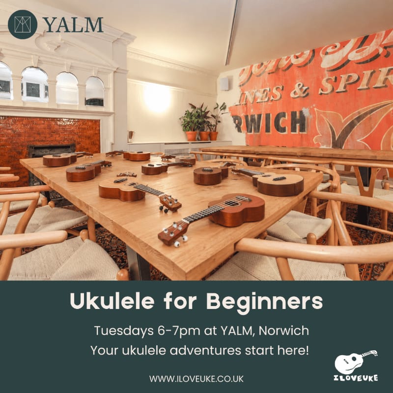 Several ukuleles are arranged on a wooden table in the foreground, with a large vintage sign and a fireplace visible in the background.