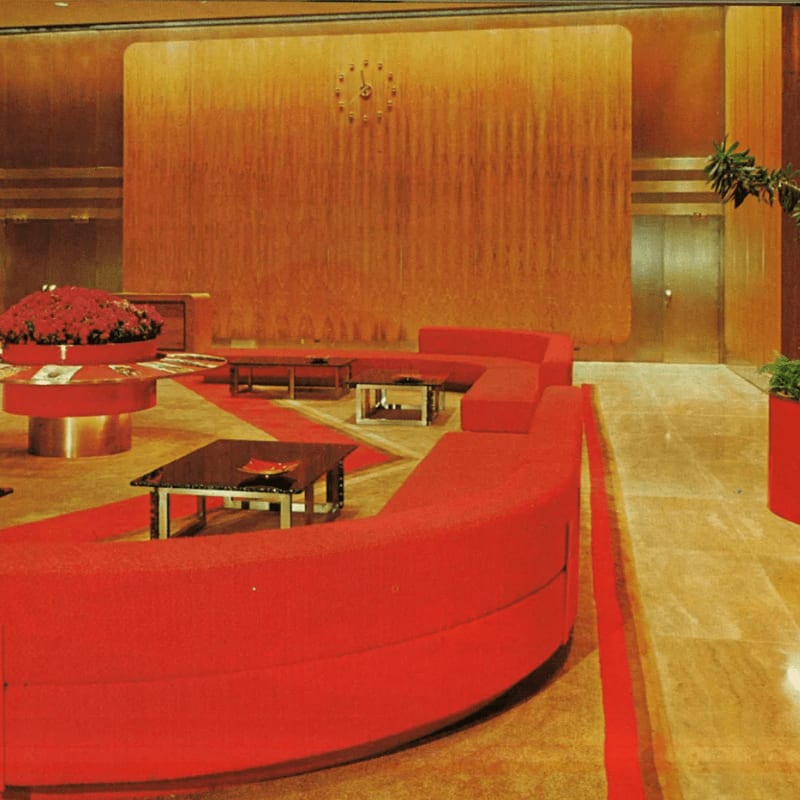 A vibrant red curved sofa dominates the foreground, leading the eye towards a large, textured orange wall with a clock in the background.