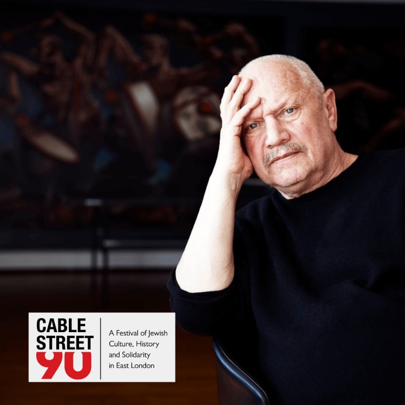 An older man with his hand on his forehead sits in front of a painting, with a "Cable Street 90" festival logo in the lower left corner.