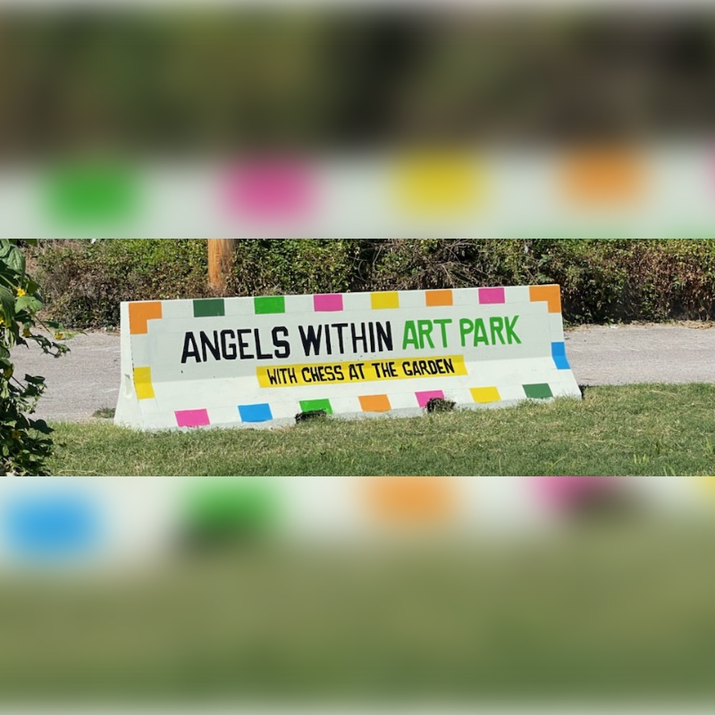 A white concrete barrier with colorful squares and text "ANGELS WITHIN ART PARK WITH CHESS AT THE GARDEN" sits on green grass in front of a blurred background of trees and sky.