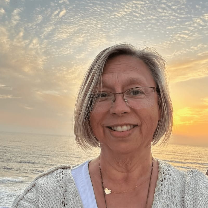 A woman with short gray hair and glasses smiles at the camera in the foreground, with a serene ocean and a dramatic sunset sky in the background.